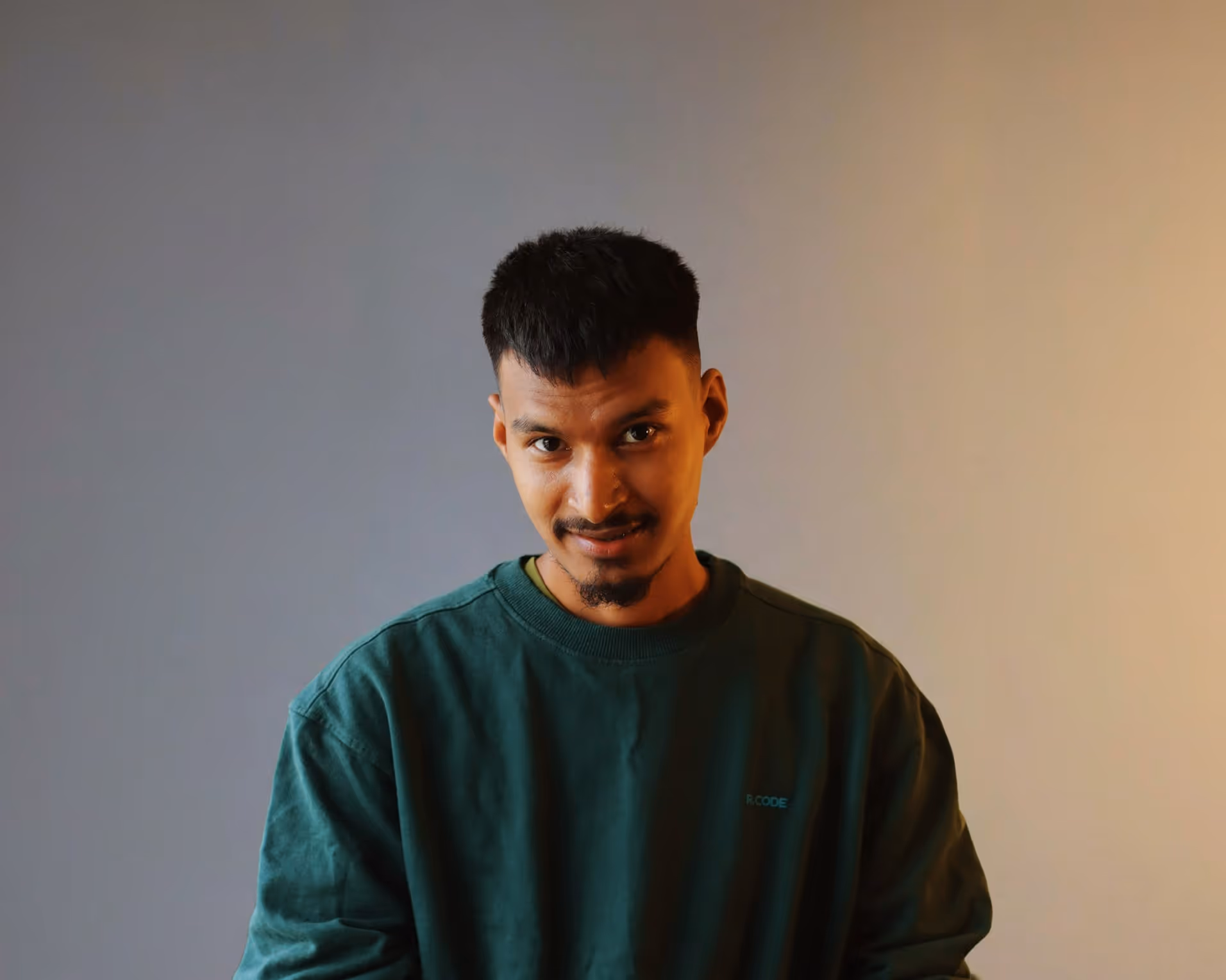 Young man with short black hair and a goatee smiling slightly, wearing a dark green sweatshirt against a two-tone gray and beige background.