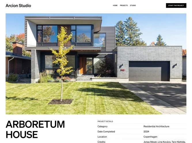 Modern two-story house with gray brick exterior, large windows, a black garage door, and a young tree planted in the front lawn.