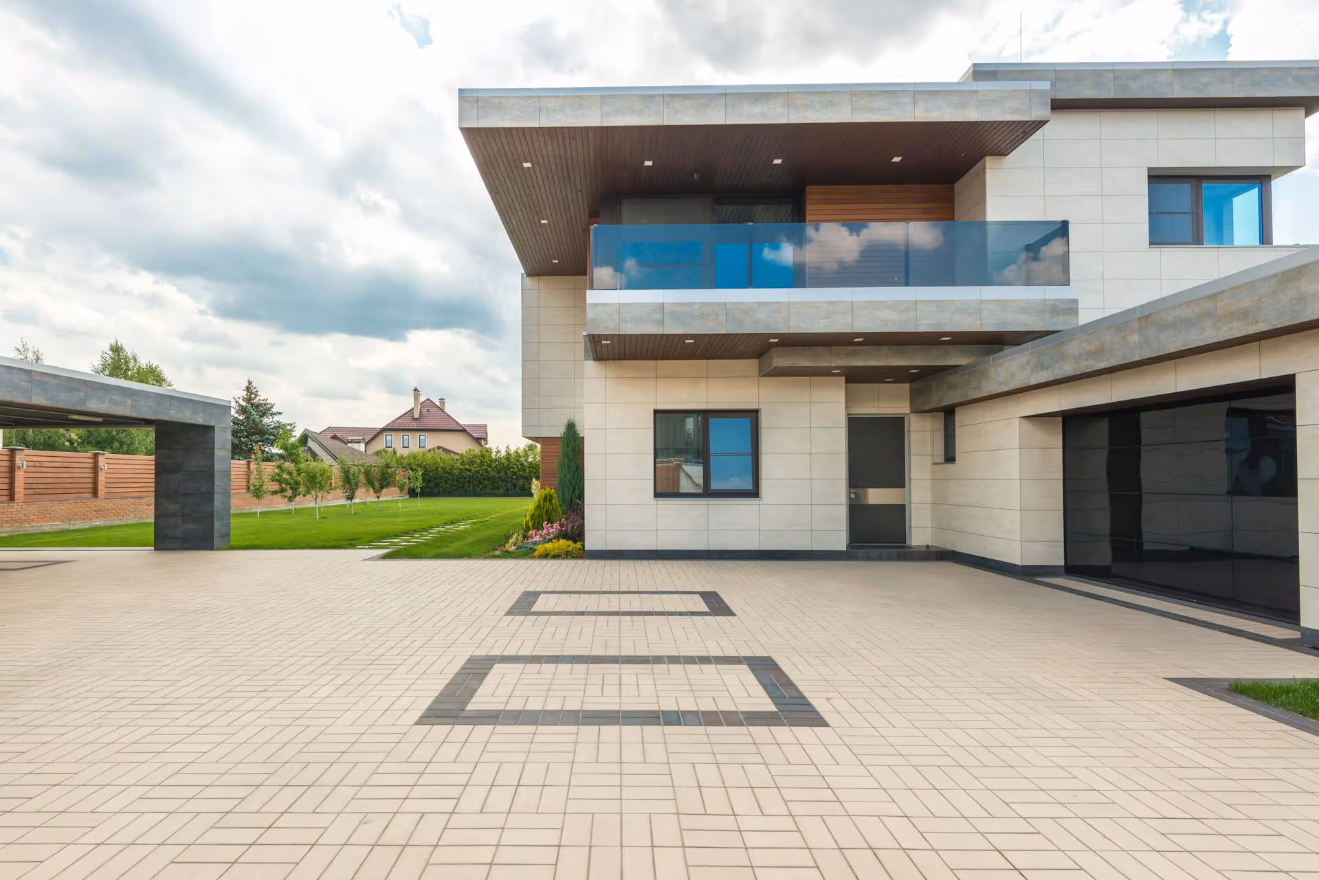Modern two-story house with large tiled driveway, glass balcony, and adjacent green lawn with garden.