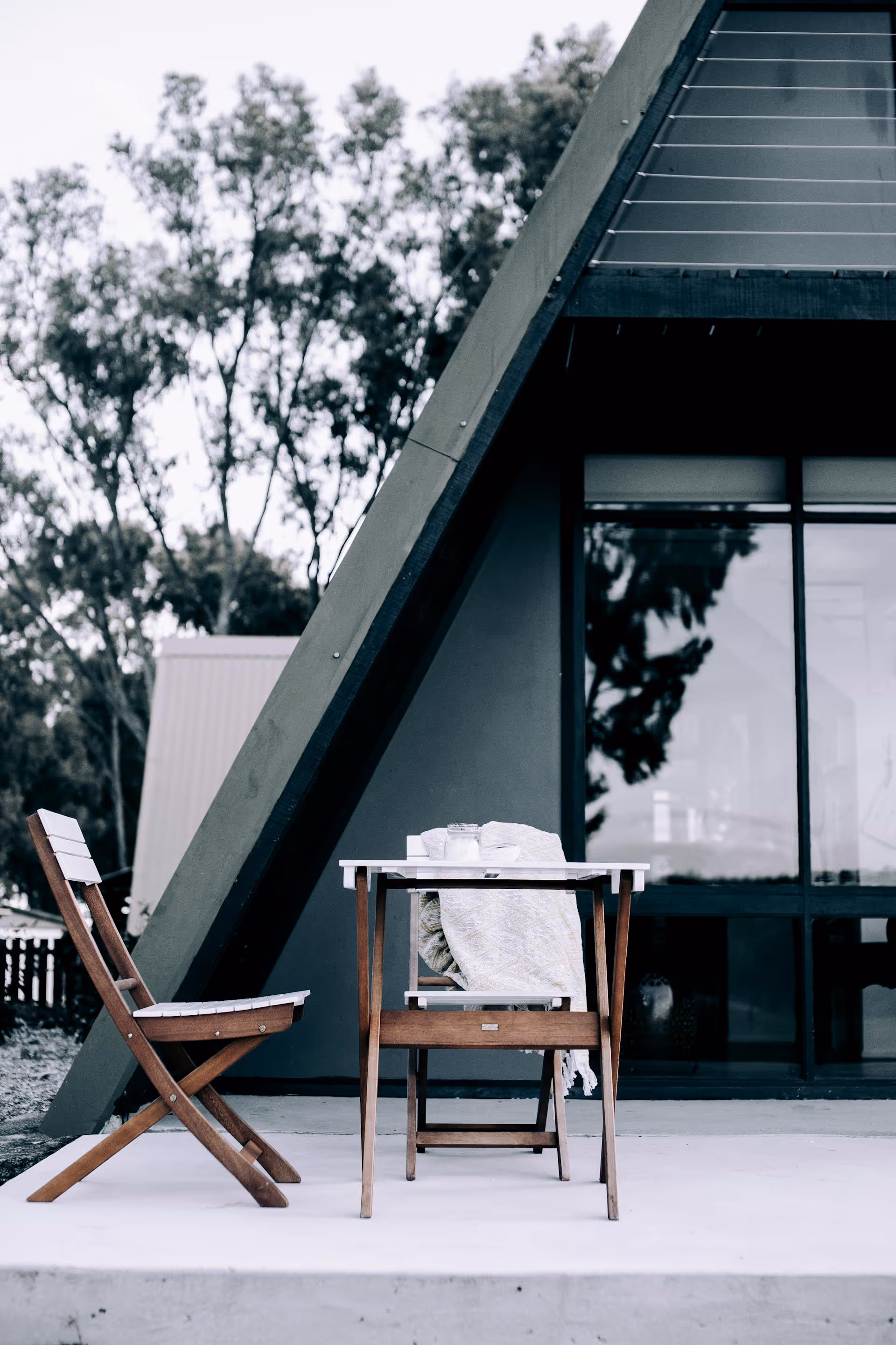 Wooden table and two chairs on a porch in front of an A-frame house with large glass windows, one chair draped with a light-colored blanket.
